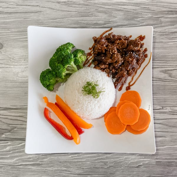 Teriyaki beef with white rice, broccoli, red and orange bell peppers, and sliced carrots on a white plate.