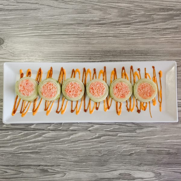 Six sushi rolls with pink filling and green wraps, drizzled with orange sauce on a white plate.